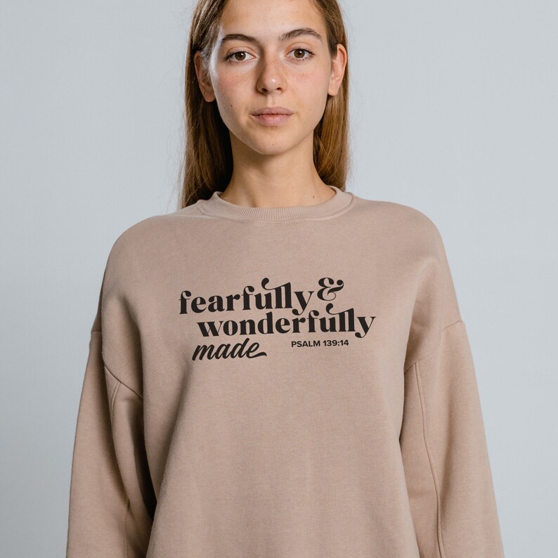 Pu&ograve; includere: Una felpa color marrone chiaro con il testo "fearfully & wonderfully made" in lettere nere. Il testo &egrave; in uno stile di font retr&ograve;. Il testo "PSALM 139:14" &egrave; stampato in lettere nere pi&ugrave; piccole sotto il testo principale.