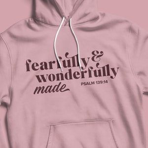 Pu&ograve; includere: Una felpa con cappuccio rosa con il testo "fearfully & wonderfully made" in lettere marroni e il riferimento biblico "PSALM 139:14" in lettere marroni pi&ugrave; piccole.