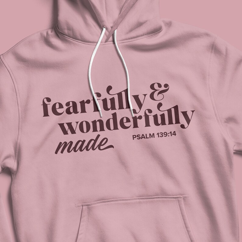 Pu&ograve; includere: Una felpa con cappuccio rosa con il testo "fearfully & wonderfully made" in lettere marroni e il riferimento biblico "PSALM 139:14" in lettere marroni pi&ugrave; piccole.