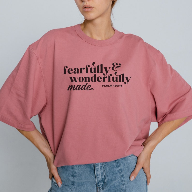 Pu&ograve; includere: Una maglietta rosa con il testo "fearfully & wonderfully made" in lettere nere. Il testo "PSALM 139:14" &egrave; anche stampato in nero sotto il testo principale.