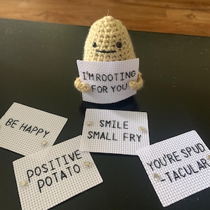 May include: A crocheted potato holding a sign that says "I'm rooting for you". There are four other signs that say "Be Happy", "Positive Potato", "Smile Small Fry", and "You're Spud-tacular".