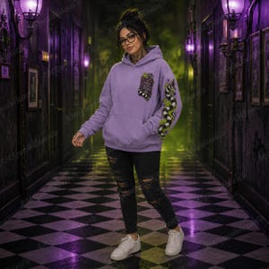 Puede incluir: Una sudadera con capucha de color lavanda que presenta un gr&aacute;fico de un libro y una serpiente a rayas blancas y negras en la manga izquierda. La sudadera tiene un bolsillo tipo canguro y se combina con vaqueros negros desgastados y zapatillas blancas.