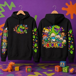 Puede incluir: Una sudadera negra con capucha que presenta un gráfico de los personajes de Rugrats y un dinosaurio verde en la parte posterior, con un gráfico de dinosaurio más pequeño en el frente. Las mangas están decoradas con huellas verdes, lápices y la palabra Rugrats.
