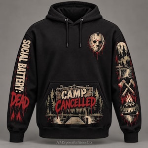 May include: A black hooded sweatshirt featuring a hockey mask graphic on the chest, the text "CAMP CANCELLED" on a wooden sign, "SOCIAL BATTERY: DEAD" on the left sleeve, and a forest, axes, and campfire scene on the right sleeve.