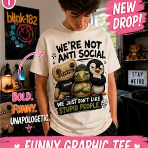 May include: Cream-colored graphic tee with the text "WE'RE NOT ANTI SOCIAL" and three cartoon characters. The shirt also says "WE JUST DON'T LIKE STUPID PEOPLE." A "blink-182" poster is visible in the background. The shirt is part of a "NEW DROP!" collection.