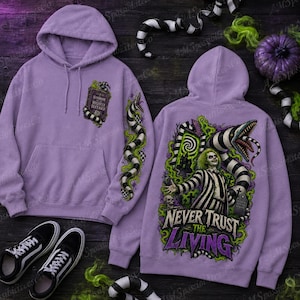 Puede incluir: Una sudadera con capucha de color morado claro que presenta un gr&aacute;fico frontal de un libro titulado Handbook for the Recently Deceased y un gr&aacute;fico trasero de un personaje con traje a rayas con el texto Never Trust the Living rodeado por una serpiente.