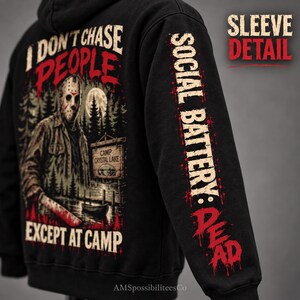 May include: A black hooded sweatshirt featuring a graphic of a hockey-masked figure holding a machete near a Camp Crystal Lake sign. The text reads I DON'T CHASE PEOPLE EXCEPT AT CAMP. The sleeve features the text SOCIAL BATTERY: DEAD.