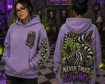 May include: A lavender hooded sweatshirt featuring a graphic of the Handbook for the Recently Deceased on the front. The back displays a Beetlejuice character illustration with a sandworm, a graveyard, and the text Never Trust the Living.