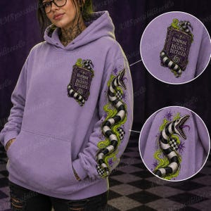 Puede incluir: Una sudadera con capucha de color morado claro que presenta un libro titulado Handbook for the Recently Deceased en el pecho y una serpiente a rayas blancas y negras con humo verde y estrellas moradas en la manga.