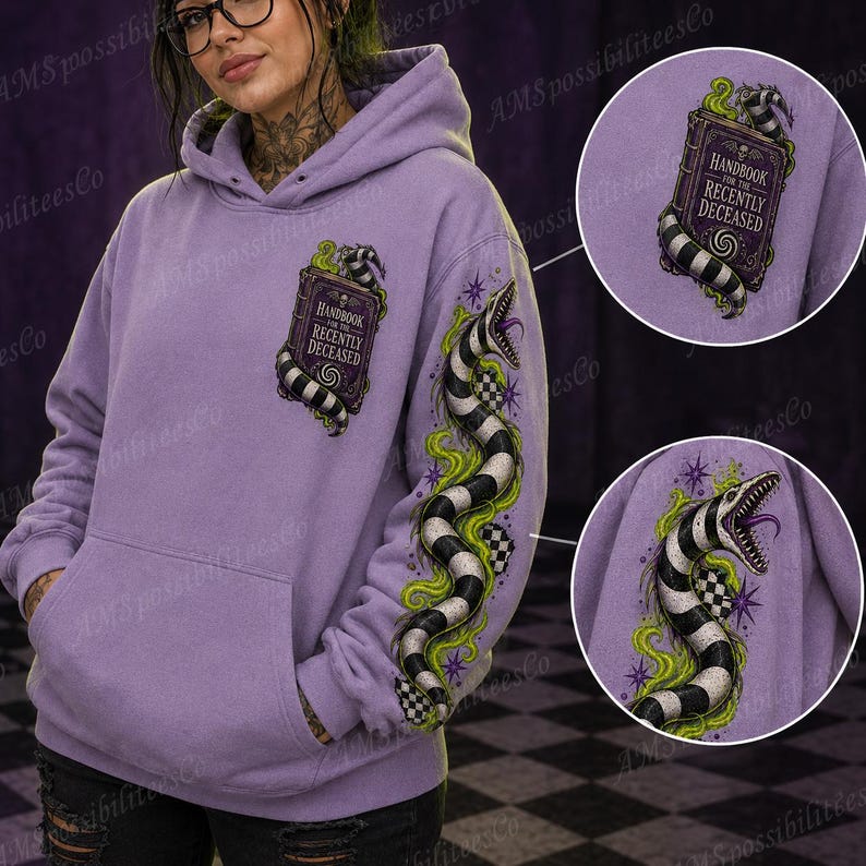 Puede incluir: Una sudadera con capucha de color morado claro que presenta un libro titulado Handbook for the Recently Deceased en el pecho y una serpiente a rayas blancas y negras con humo verde y estrellas moradas en la manga.