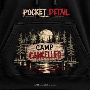 May include: A black hoodie featuring a graphic print of a wooden sign that reads CAMP CANCELLED in red letters. The design includes a forest of pine trees, a full moon, and a reflection in water. The text POCKET DETAIL is printed above the pocket.