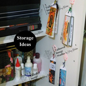 May include: A white shelf with various items, including bottles of liquid, a stack of black notebooks, and several colorful bookmarks hanging from hooks. The bookmarks are labeled "A Tailored Image Studio by Stacy Bledsoe" and feature different designs, including a butterfly, a flower, and a cityscape. The text "Storage Ideas" is written in black on a white background.