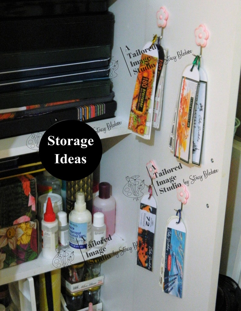May include: A white shelf with various items, including bottles of liquid, a stack of black notebooks, and several colorful bookmarks hanging from hooks. The bookmarks are labeled "A Tailored Image Studio by Stacy Bledsoe" and feature different designs, including a butterfly, a flower, and a cityscape. The text "Storage Ideas" is written in black on a white background.