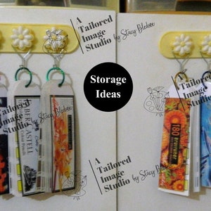 May include: A white wall with two rows of colorful, hanging, labeled, storage containers. The containers are labeled "Tailored Image Studio" and "180 Brutruner". The containers are hanging from a yellow bar with three white daisy-shaped hooks.