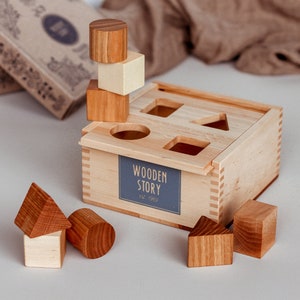 Può includere: Giocattolo di smistamento di forme in legno con finitura in legno chiaro. Lo smistatore ha un'etichetta blu con la scritta "Wooden Story Est. 1969". Include varie forme di legno in tonalità di legno naturale, come cubi, cilindri e triangoli.