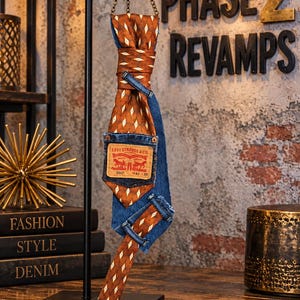 May include: A necktie crafted from a combination of brown fabric with a white diamond pattern and blue denim fabric. The tie features a prominent Levi Strauss & Co. leather patch on a denim pocket detail, suspended from a black metal display stand.