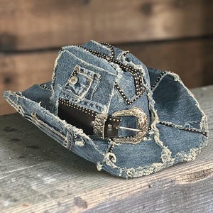 May include: A denim cowboy hat with a distressed look. The hat features a black leather band with a silver buckle and is accented with black and white polka dot trim. The hat has a small pocket on the front.