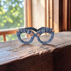 May include: A pair of handcrafted, dark blue eyeglasses with a unique, textured design. The frames are wrapped in a dark blue material, and the lenses are connected with small, copper-colored rivets. The glasses are displayed on a weathered wooden surface.
