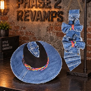 May include: A black hat with a wide blue denim brim and a braided multi-coloured band with a brass buckle, accompanied by a hanging denim necktie featuring two denim bows with braided multi-coloured centres and frayed edges.