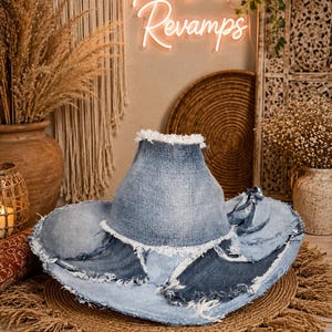 May include: A wide-brimmed hat constructed from repurposed light and medium blue denim fabric. The hat features a high crown and a broad, uneven brim with frayed, raw edges throughout. The denim panels are stitched together in a patchwork style.