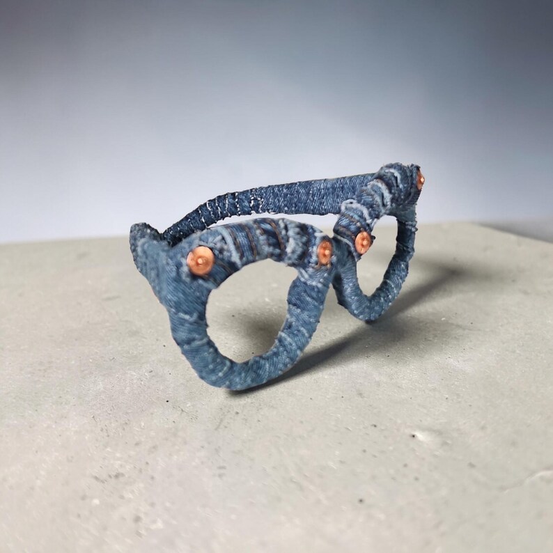 May include: A pair of eyeglasses crafted from denim fabric. The frames are a faded blue with copper-colored rivets at the joints. The glasses are set against a neutral background, highlighting the unique texture and design.