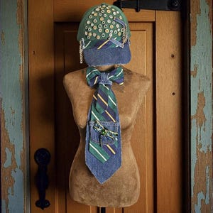May include: A denim baseball cap and tie set displayed on a mannequin. The green cap is embellished with small gold-colored accents and chains. The tie and bow tie feature green, blue, and yellow stripes, with a small pocket on the tie.