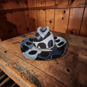 May include: A cowboy hat featuring a black base with layered patches of light and dark blue denim fabric in a circular pattern. The hat includes a decorative band around the crown adorned with small, reflective silver-toned studs or beads.
