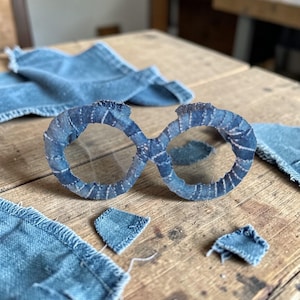 May include: A pair of eyeglasses made from denim fabric, with a unique infinity symbol design. The glasses are a light blue color, with visible stitching details. Several denim fabric scraps are scattered around the glasses on a wooden surface.