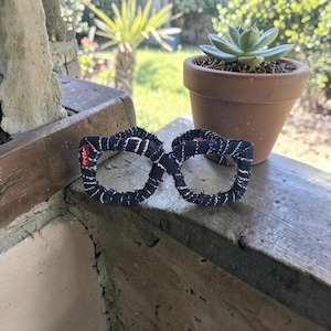 Puede incluir: Un par de gafas con estampado azul oscuro y blanco con el logo de Levi's en el lado izquierdo. Las gafas están hechas de una tela texturizada y descansan sobre una superficie de madera desgastada. Una pequeña planta suculenta en maceta está en el fondo.