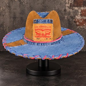 May include: A wide-brimmed hat constructed from blue denim and brown suede panels. The front features a tan Levi Strauss & Co. patch with red text and graphics. The brim is accented with bright pink stitching along the edge and a pink band.