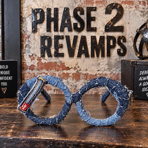 May include: A pair of circular decorative glasses frames wrapped in various shades of blue denim fabric. The frames feature frayed edges, visible stitching, and a small red fabric tag on the left side. The design mimics the shape of eyeglasses.