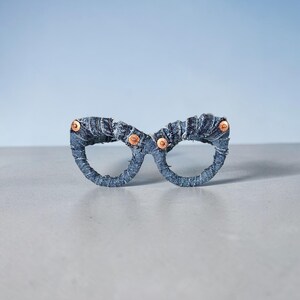 May include: A pair of eyeglasses crafted from denim fabric. The frames are a dark blue, with copper-colored rivets at the temples. The glasses are set against a light blue background, showcasing a unique, handcrafted accessory.