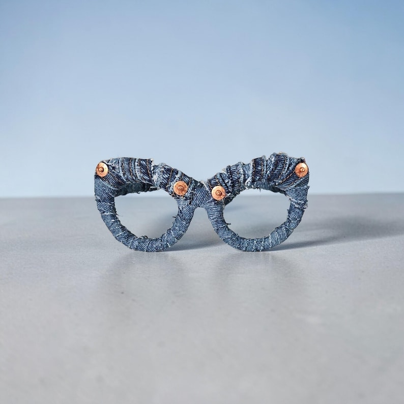 May include: A pair of eyeglasses crafted from denim fabric. The frames are a dark blue, with copper-colored rivets at the temples. The glasses are set against a light blue background, showcasing a unique, handcrafted accessory.