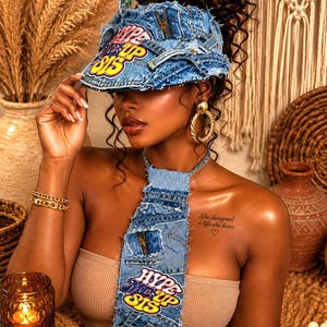 Può includere: Una visiera in denim blu e una cravatta in denim abbinata, entrambe con bordi sfrangiati e toppe con la scritta HYPE ME UP SIS in giallo, viola e bianco. La visiera presenta un dettaglio con cerniera metallica sulla parte superiore.