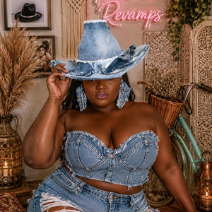 May include: A denim outfit consisting of a strapless corset-style top with frayed edges and matching distressed denim shorts. The ensemble is paired with a wide-brimmed denim hat featuring a frayed, layered brim and matching denim earrings.