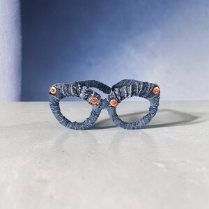 May include: A pair of eyeglasses crafted from blue denim fabric. The frames are wrapped in the denim material and feature copper-colored rivets at the temples. The glasses are set against a blue and white background.
