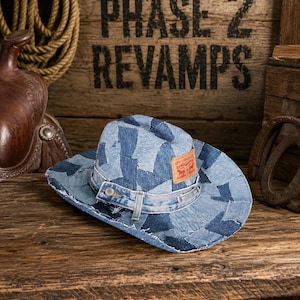 May include: A cowboy hat constructed from various shades of blue denim fabric patches. The hat features a denim band with a metal button and a tan leather Levi Strauss brand patch on the front. The edges of the brim have a frayed appearance.