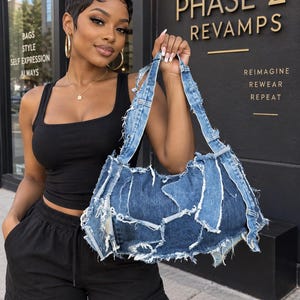 May include: A medium-sized shoulder bag constructed from repurposed blue denim fabric. The bag features a patchwork design with multiple frayed, raw-edge denim panels stitched together, including visible belt loops and seams, with two denim straps.