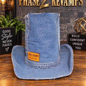 May include: A blue denim cowboy hat with a tall, cylindrical crown and a wide, curved brim. The hat features frayed edges and a rectangular tan leather patch on the front with the text Phase 2 Revamps One of a Kind.