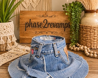 May include: A wide-brimmed hat constructed from light blue denim fabric, featuring a waistband with belt loops, a red brand tag, and a tan leather patch. The brim is composed of layered denim panels with frayed, raw edges throughout the design.
