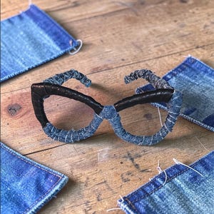 Op de afbeelding: Handgemaakte bril met een met denim omwikkelde montuur en donkerbruine veren. De bril is omgeven door kleine denim stoffen vierkanten op een houten oppervlak. Het montuur is een uniek, artistiek ontwerp.