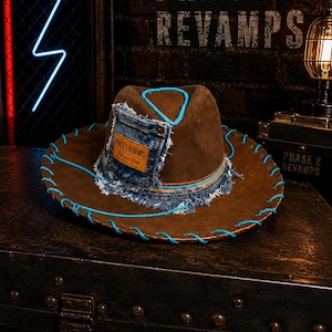 May include: A brown hat featuring a blue denim pocket patch with frayed edges and a leather label reading PHASE 2 REVAMPS. The hat is accented with bright blue cord stitching along the brim and crown, creating a distinct contrast against the brown.