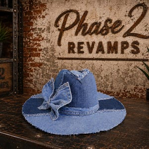 Puede incluir: Un sombrero de ala ancha confeccionado con varios tonos de tela vaquera azul. El sombrero presenta un lazo de mezclilla prominente unido al costado y muestra bordes deshilachados a lo largo del ala y la copa, con un diseño de parches.