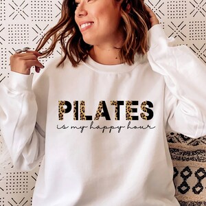 May include: White sweatshirt with the text "PILATES is my happy hour" in black with a leopard print pattern.