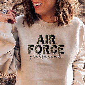 May include: A beige sweatshirt with the words "AIR FORCE girlfriend" printed on the front. The word "FORCE" is in a camouflage pattern.