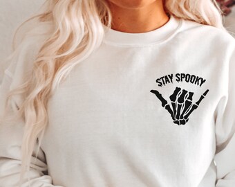 Może przedstawiać: Biała bluza z czarnym szkieletowym ręką wykonującą znak shaka i tekstem "Stay Spooky" z przodu.
