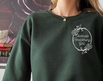 Puede incluir: Una sudadera verde oscuro con un diseño gráfico blanco que dice "Obstinate Headstrong Girl" en un círculo con acentos florales.