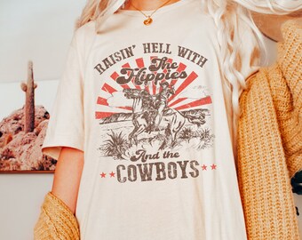 Könnte beinhalten: Ein weißes T-Shirt mit einem Vintage-Grafikdruck, auf dem "Raisin' Hell With The Hippies And The Cowboys" steht. Die Grafik zeigt eine rote Sonne mit Strahlen und zwei Cowboys, die auf Pferden reiten.