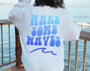 Puede incluir: Sudadera blanca con un gráfico azul degradado que dice "MAKE SOME WAVES" con una ola azul debajo.