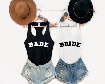 Op de afbeelding: Twee tanktops, een zwarte en een witte, met de woorden "BABE" en "BRIDE" gedrukt in wit op de zwarte tanktop en in zwart op de witte tanktop. De tanktops hangen aan hangers met bruine en zwarte hoeden erboven.